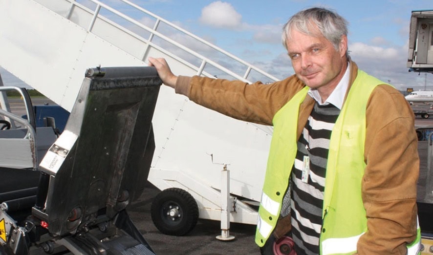 A man wearing a reflective safety vest stands next to an open luggage conveyor belt and a belt loader at an airport, with airport equipment visible in the background.