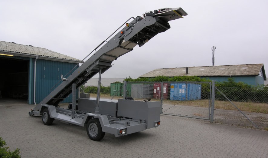 A belt loader is parked on a paved surface near a chain-link fence, with industrial buildings and shipping containers in the background.