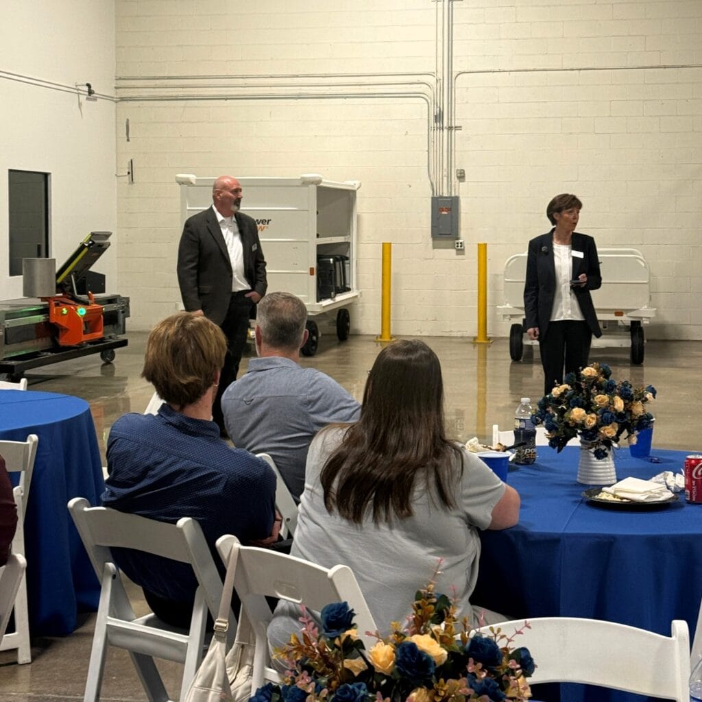 Two presenters speak to a seated audience at a corporate event in an industrial warehouse setting, with Power Stow equipment visible in the background.